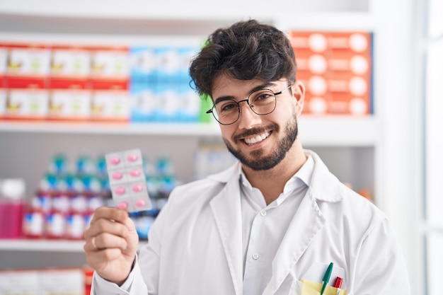 Jeune homme hispanique pharmacien souriant confiant tenant des pilules à la pharmacie