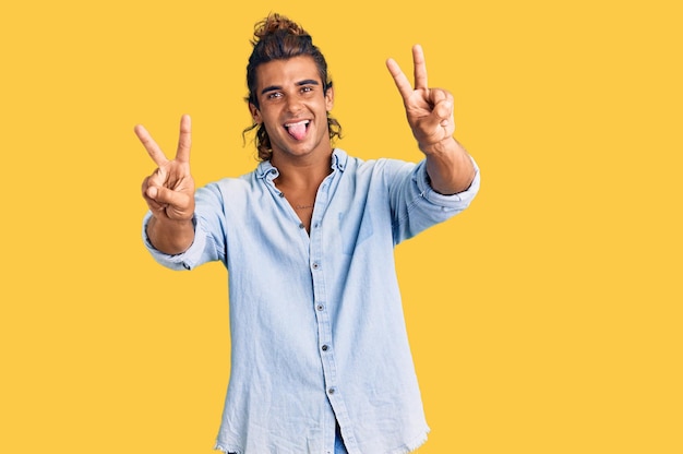 Jeune homme hispanique portant un style d'été souriant avec la langue montrant les doigts des deux mains faisant le signe de la victoire. numéro deux.