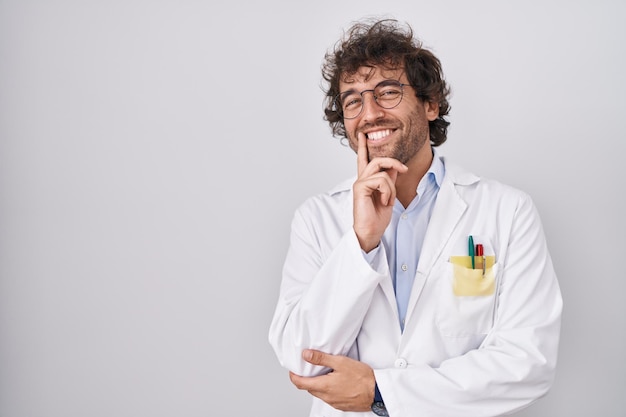 Un jeune homme hispanique portant un uniforme de médecin a l'air confiant devant la caméra en souriant les bras croisés et la main levée sur le menton. penser positivement.