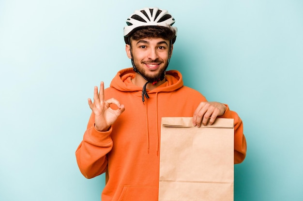 Jeune homme hispanique portant un vélo casque tenant un plat à emporter isolé sur fond bleu joyeux et confiant montrant un geste ok