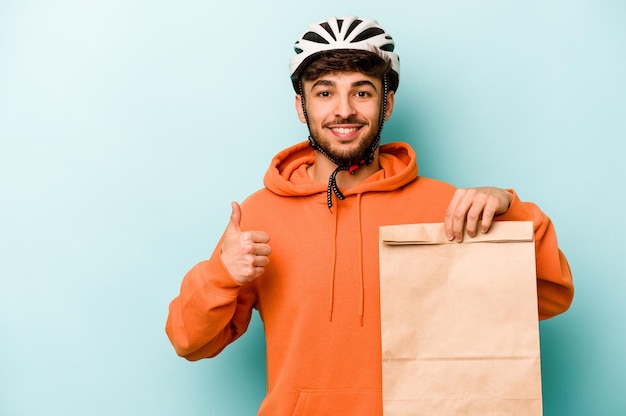 Jeune homme hispanique portant un vélo casque tenant un plat à emporter isolé sur fond bleu souriant et levant le pouce vers le haut