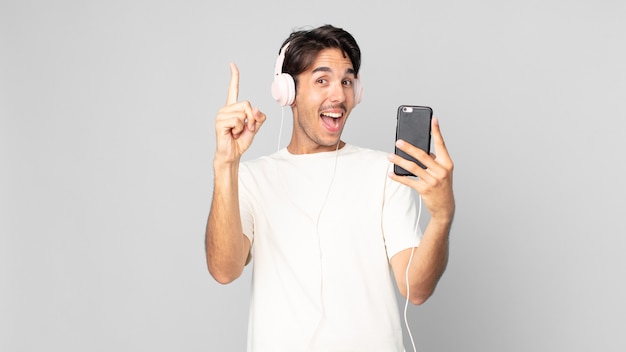 Jeune homme hispanique se sentant comme un génie heureux et excité après avoir réalisé une idée avec des écouteurs et un smartphone