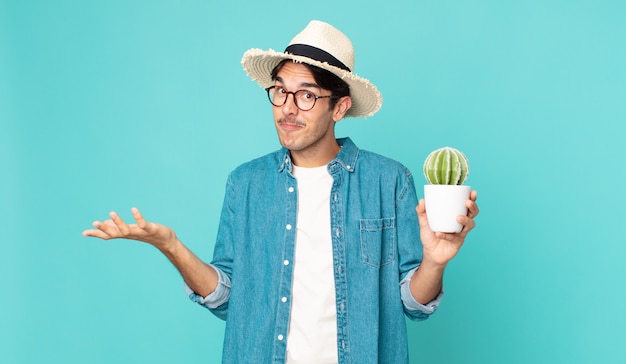 Jeune homme hispanique se sentant perplexe et confus et doutant et tenant un cactus