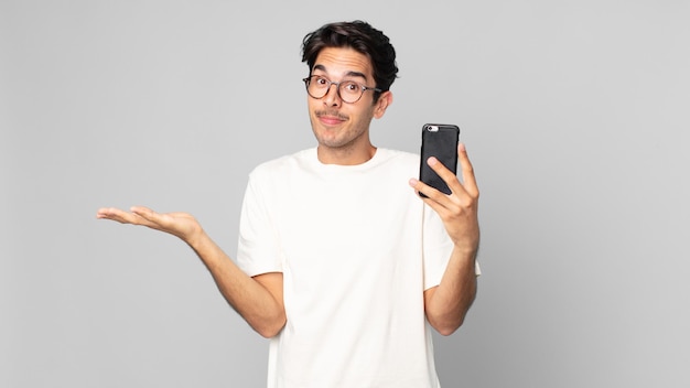 Jeune homme hispanique se sentant perplexe et confus et doutant et tenant un smartphone