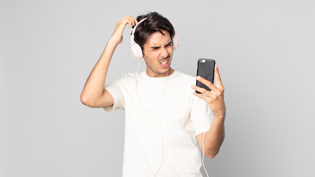 Jeune homme hispanique se sentant perplexe et confus, se grattant la tête avec des écouteurs et un smartphone