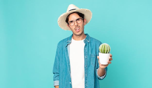 Jeune homme hispanique se sentant perplexe et confus et tenant un cactus