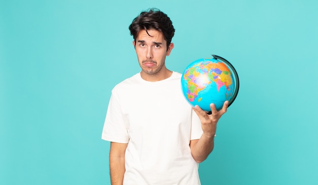 Jeune homme hispanique se sentant triste et pleurnichard avec un regard malheureux et pleurant et tenant une carte du globe terrestre