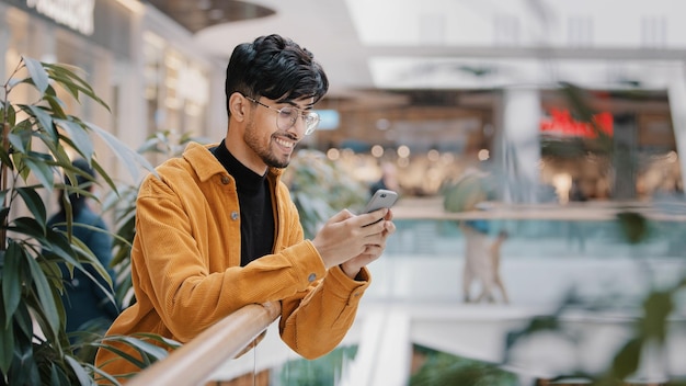 Jeune homme hispanique se tient dans un centre commercial tenant un téléphone souriant envoyant un e-mail sur le réseau social