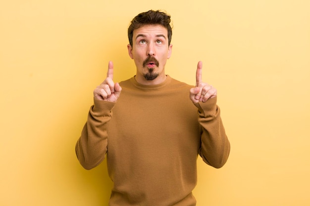 Jeune homme hispanique semblant choqué étonné et bouche bée pointant vers le haut avec les deux mains pour copier l'espace