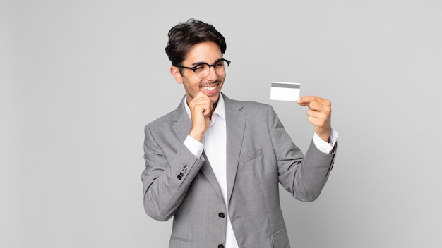 Jeune homme hispanique souriant avec une expression heureuse et confiante avec la main sur le menton et tenant une carte de crédit