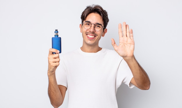 Jeune homme hispanique souriant joyeusement, agitant la main, vous accueillant et vous saluant. concept de vaporisateur de fumée