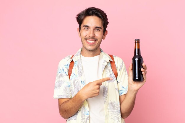 Jeune homme hispanique souriant joyeusement, se sentant heureux et pointant vers le côté et tenant une bouteille de bière