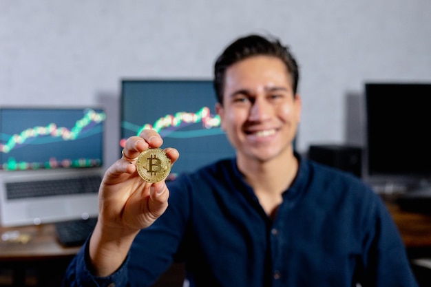 Jeune homme hispanique souriant montrant une pièce de monnaie en bitcoin. ordinateur avec. Ordinateurs affichant des courbes de croissance de la devise de valeur en arrière-plan.