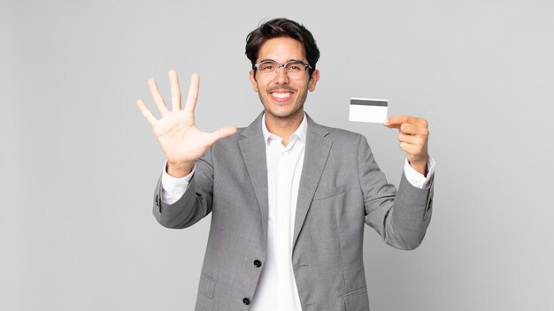 Jeune homme hispanique souriant et semblant amical, montrant le numéro cinq et tenant une carte de crédit