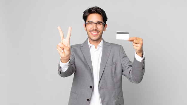 Jeune homme hispanique souriant et semblant amical, montrant le numéro deux et tenant une carte de crédit