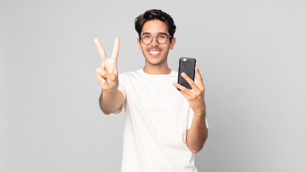 Jeune homme hispanique souriant et semblant amical, montrant le numéro deux et tenant un smartphone