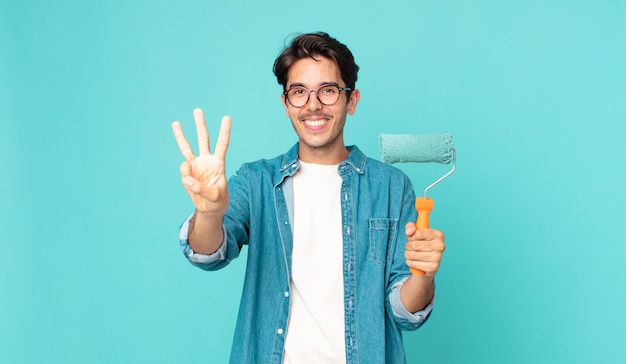 Jeune homme hispanique souriant et semblant amical, montrant le numéro trois et tenant un rouleau à peinture