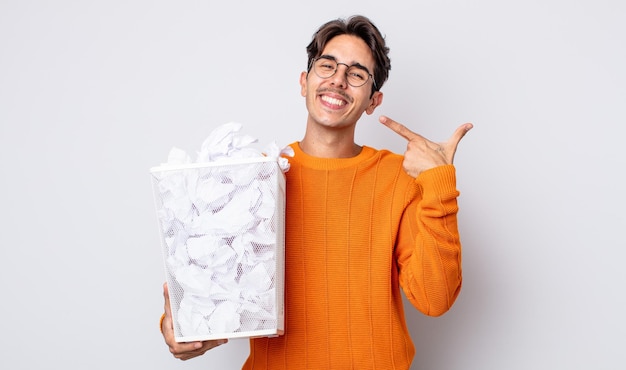 Jeune homme hispanique souriant en toute confiance pointant vers son large sourire. concept de corbeille de boules de papier