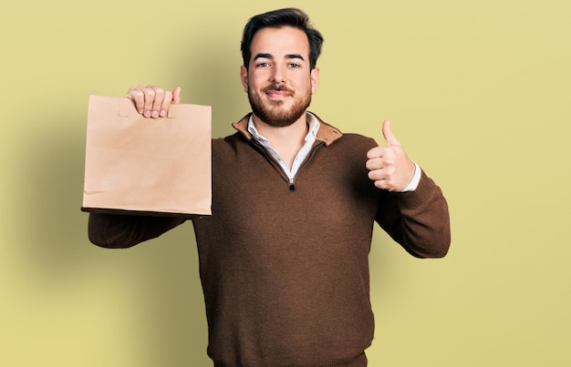 Jeune homme hispanique tenant un sac en papier à emporter souriant heureux et positif, pouce vers le haut faisant un excellent signe d'approbation