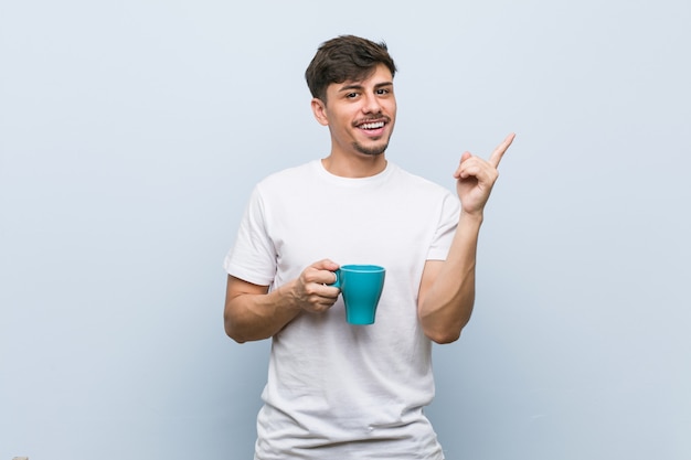 Jeune homme hispanique tenant une tasse en souriant joyeusement pointant avec l'index loin.