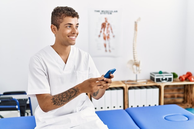 Jeune homme hispanique travaillant comme physiothérapeute utilisant un smartphone dans la salle de physiothérapie