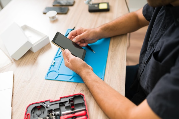 Jeune homme hispanique utilisant des outils pour ouvrir un smartphone endommagé. Technicien masculin réparant un écran cassé à l'atelier de réparation