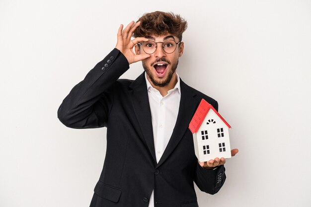 Jeune homme immobilier arabe tenant une maison modèle isolée sur fond isolé excité en gardant le geste ok sur les yeux.