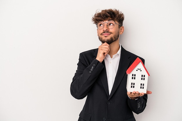 Jeune homme immobilier arabe tenant une maison modèle isolée sur fond isolé regardant de côté avec une expression douteuse et sceptique.