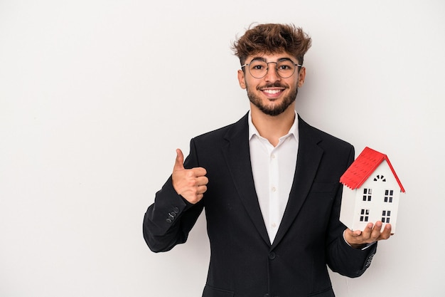 Jeune homme immobilier arabe tenant une maison modèle isolée sur fond isolé souriant et levant le pouce vers le haut