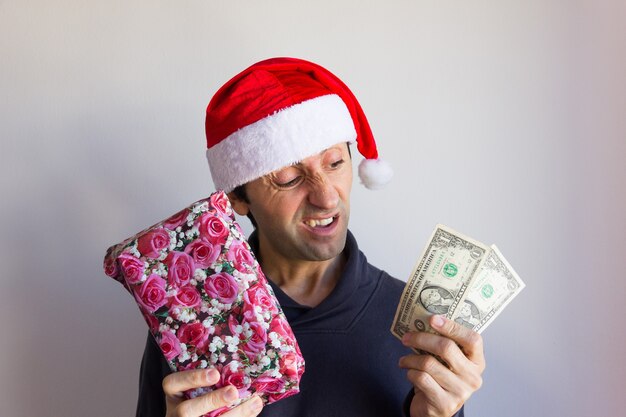 Jeune homme incrédule avec chapeau de Père Noël tenant un cadeau emballé et peu d'argent avec un regard sceptique