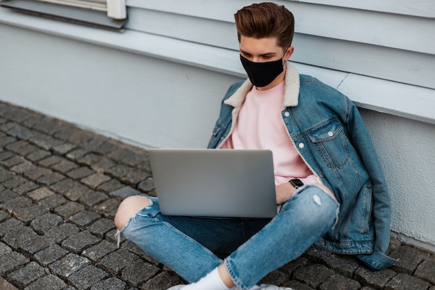 Jeune homme indépendant à la mode en masque médical noir dans des vêtements de jeans décontractés en denim élégant est assis avec un ordinateur portable et travaille à distance sur un projet créatif. Le créateur à la mode tape sur le clavier à l'extérieur.