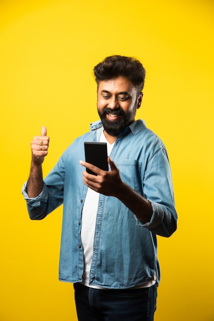 Jeune homme indien barbu utilisant un smartphone, souriant en appelant ou en discutant avec un ami, debout sur du jaune