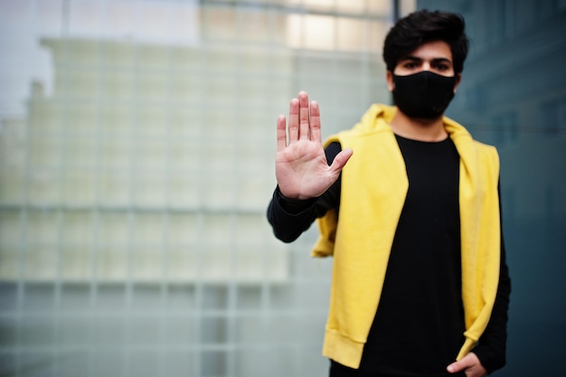 Jeune homme indien hipster urbain dans un sweat-shirt jaune à la mode Un gars sud-asiatique cool porte un sweat à capuche et un masque de protection du visage noir pendant le nouveau spectacle normal s'arrête à la main