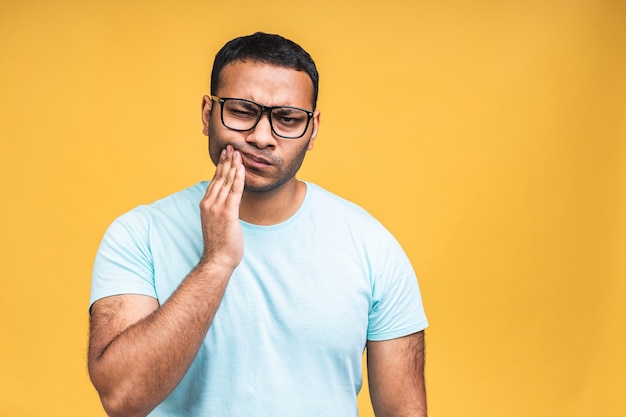 Jeune homme indien portant un t-shirt debout sur fond jaune isolé touchant la bouche avec la main avec une expression douloureuse à cause d'un mal de dents ou d'une maladie dentaire sur les dents. Notion de dentiste.