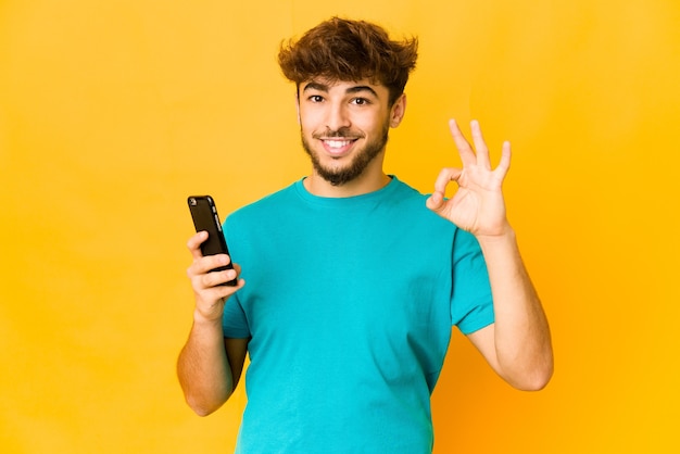 Jeune homme indien tenant un téléphone joyeux et confiant montrant un geste correct.