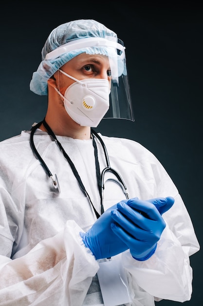 Jeune homme infirmier hospitalier en masque de protection médicale, gants et vêtements de protection isolés sur fond sombre.