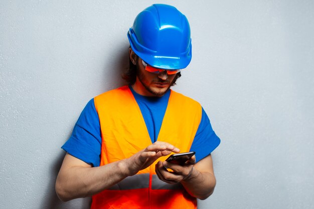 Jeune homme ingénieur ouvrier du bâtiment, tapant sur smartphone, portant un équipement de sécurité sur fond texturé gris.