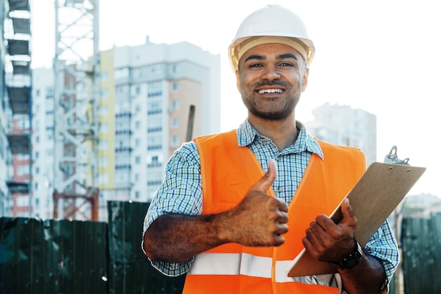 Jeune homme ingénieur en vêtements de travail debout sur chantier avec presse-papiers