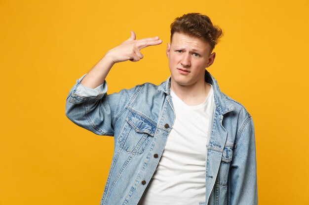 Un jeune homme insatisfait en vêtements décontractés en denim pointe les doigts vers la tête comme s'il se tirait dessus isolé sur fond orange jaune. Concept de mode de vie des émotions sincères des gens. Maquette de l'espace de copie.