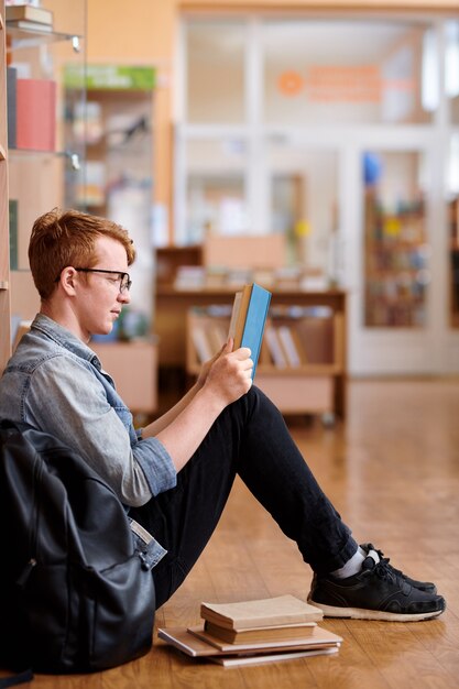 Jeune homme intelligent dans un livre de lecture de vêtements décontractés alors qu'il était assis sur le sol de la bibliothèque après les cours