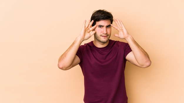 Jeune homme isolé sur beige recevant une agréable surprise, excité et levant les mains.