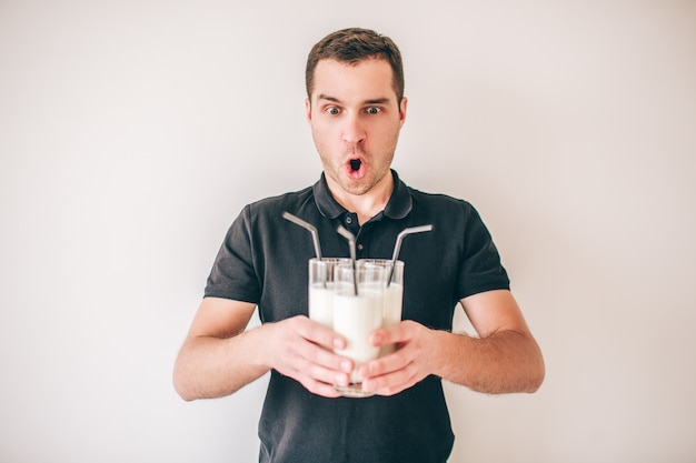 Jeune homme isolé sur blanc. Le gars étonné et étonné tient trois verres de lait frais ou de lait frappé dans les mains. Paille en plastique noir à l'intérieur.