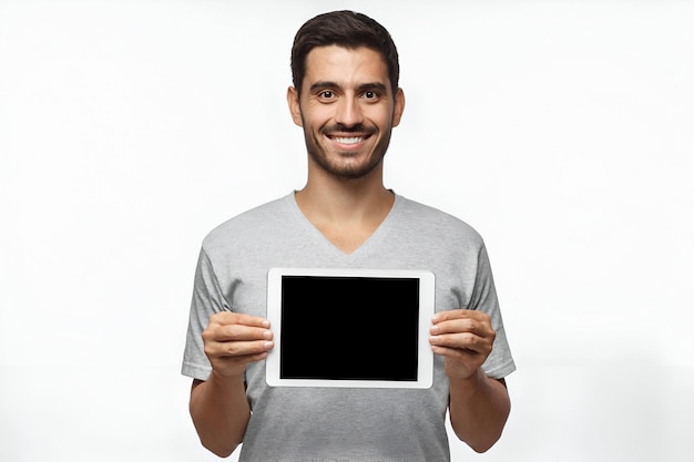 Jeune homme isolé sur un fond gris tenant une tablette et montrant un écran vide avec un sourire heureux comme si il conseillait un produit
