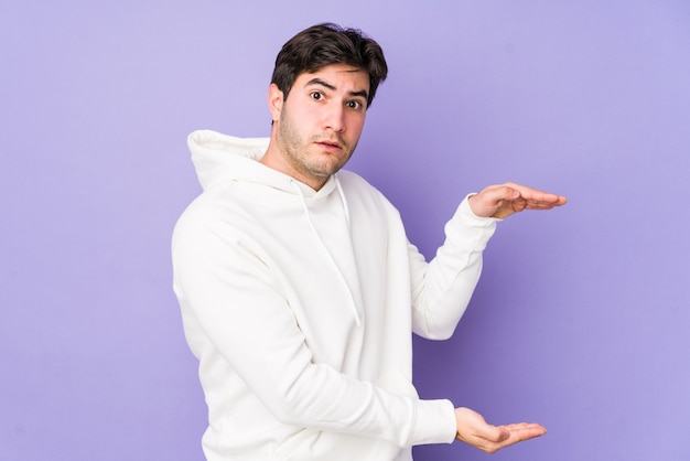 Jeune homme isolé sur fond violet choqué et étonné tenant un espace de copie entre les mains.