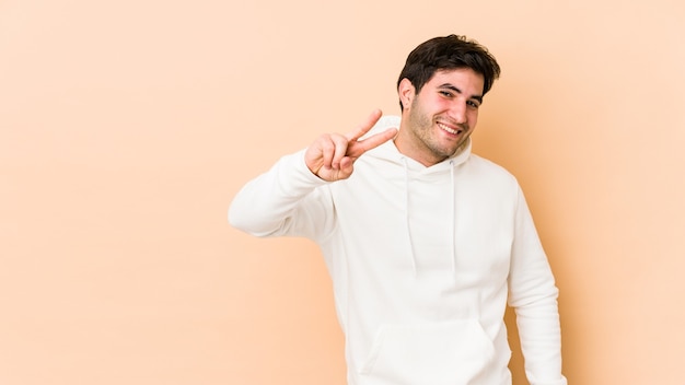 Jeune homme isolé sur un mur beige montrant le signe de la victoire et souriant largement.