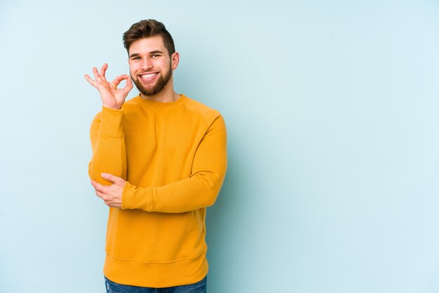 Jeune homme isolé sur le mur bleu fait un clin d'œil et détient un geste correct avec la main