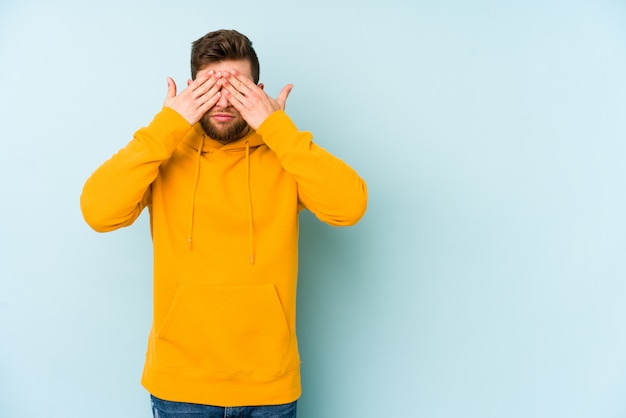 Jeune homme isolé sur le mur bleu peur couvrant les yeux avec les mains