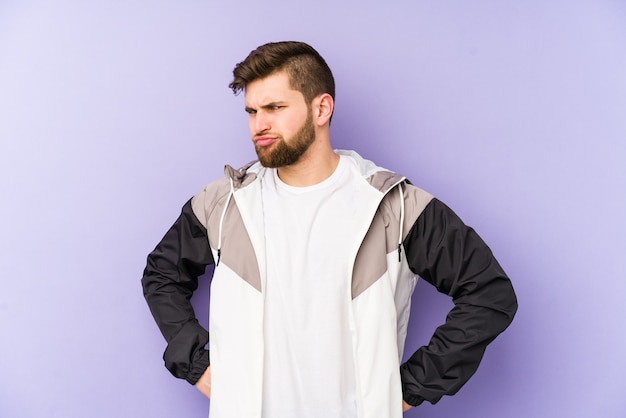 Jeune homme isolé sur un mur violet fronçant le visage de mécontentement, garde les bras croisés.