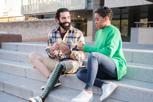 Jeune homme avec une jambe prothétique et jeune femme aux cheveux courts s'amusant à manger un sandwich à l'extérieur