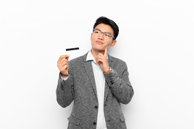 Jeune homme japonais souriant joyeusement et rêvassant ou doutant, regardant sur le côté. concept de carte de crédit.